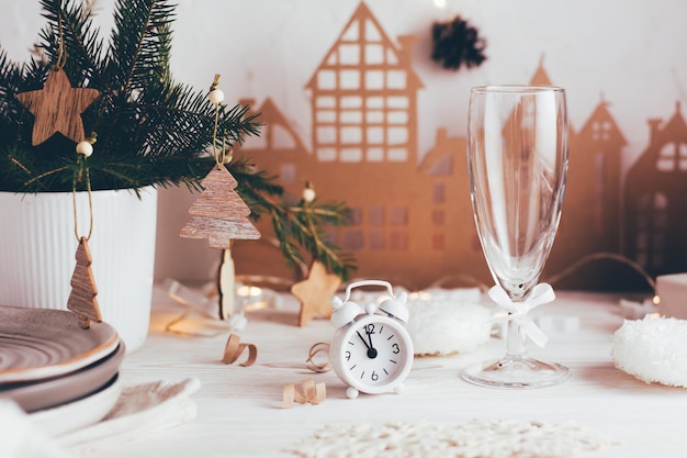 Organizzazione di una tavola natalizia festiva. Sveglia bianca e un bicchiere di champagne sullo sfondo di un bouquet di abete in un vaso e una casa di cartone. Tablescapes su tavola di legno bianco