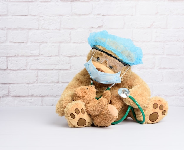 Orsacchiotto marrone si siede in occhiali protettivi di plastica, una maschera usa e getta medica e un berretto blu, concetto di pediatria