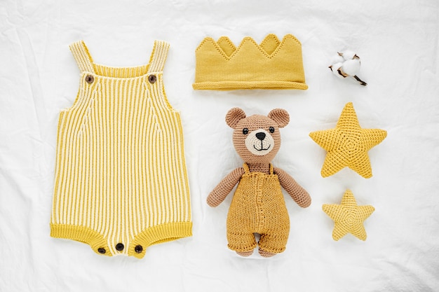 Orso giocattolo lavorato a maglia, pagliaccetto giallo e stelle giocattolo per neonato su letto bianco. Vestiti e accessori per bambini neutri di genere. Disposizione piatta, vista dall'alto