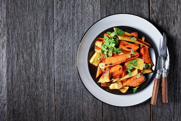 Ortaggi a radice arrostiti marocchini: pastinaca e carota con spezie ras el hanout: cannella, curcuma, anice su un piatto con coriandolo fresco e posate su uno sfondo di legno, vista dall'alto, spazio copia