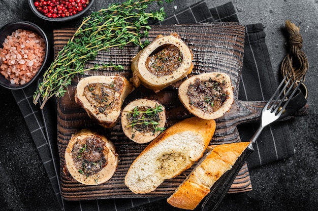 Ossa di vitello di midollo arrosto su tavola di legno con pane ed erbe aromatiche. Sfondo nero. Vista dall'alto.