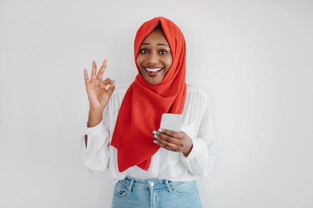 Ottima app Donna musulmana nera eccitata in hijab che utilizza lo smartphone e mostra lo sfondo della parete chiara del gesto ok