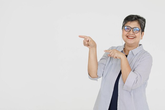 Ottimista donna asiatica di mezza età con gli occhiali che guarda la telecamera con un sorriso e che punta lontano su sfondo grigio