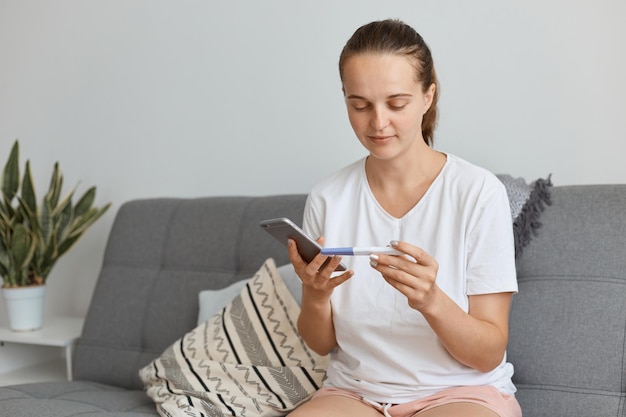 Ottimista incinta caucasica dai capelli scuri femmina che indossa abiti in stile casual con test di gravidanza in mano e tiene in mano uno smartphone, componendo il numero del marito per dirgli una buona notizia.