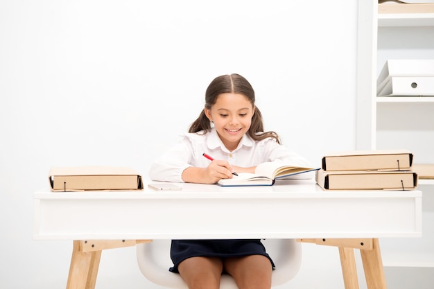 Ottimo allievo. La bambina scrive il compito mentre si siede sul tavolo sfondo bianco. Scolara che studia saggio di scrittura. La faccia felice dell'uniforme scolastica della ragazza del bambino esercita il compito domestico, copia lo spazio.