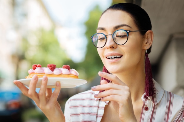 Ottimo cibo. Giovane donna allegra con gli occhiali e grandi orecchini e sorridente mentre guarda l'eclair lampone nelle sue mani