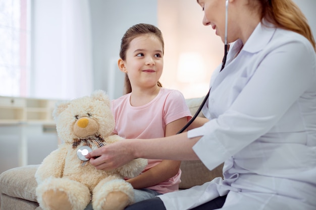 Ottimo paziente. Bambina graziosa che esamina la dottoressa che si siede accanto a lei e che cura l'orso della peluche