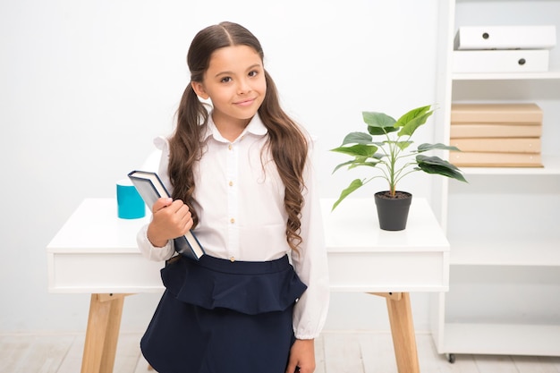 Ottimo primo ex. La bambina tiene il libro mentre sta in piedi l'interno bianco del tavolo. Scolara che studia libro di testo. Libro della tenuta della faccia felice dell'uniforme scolastica del bambino. Entusiasta della conoscenza. Pronto inserisci il primo modulo.