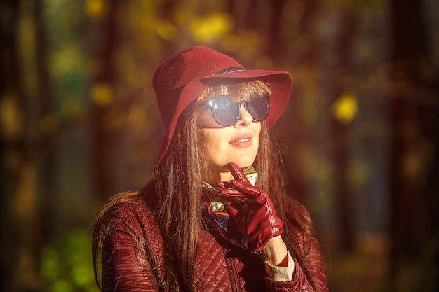 Outdoor primo piano ritratto modello di grandi dimensioni bella ragazza bruna con cappello e occhiali da sole guardando la fotocamera nella foresta d'autunno come sfondo