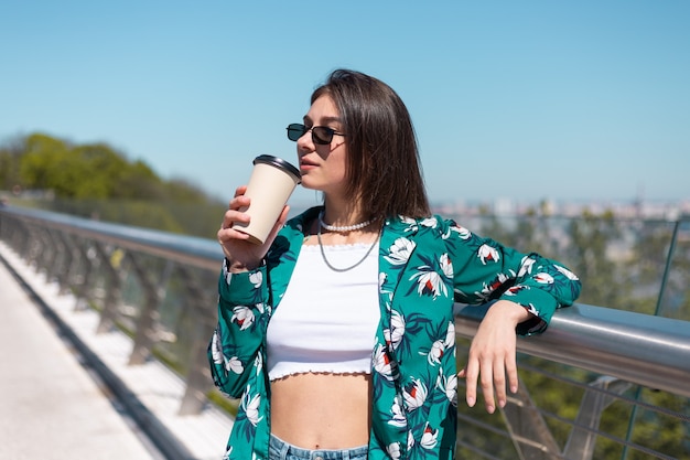 Outdoor ritratto di donna in camicia verde con una tazza di caffè che gode del sole, si trova sul ponte con vista mozzafiato sulla città al mattino