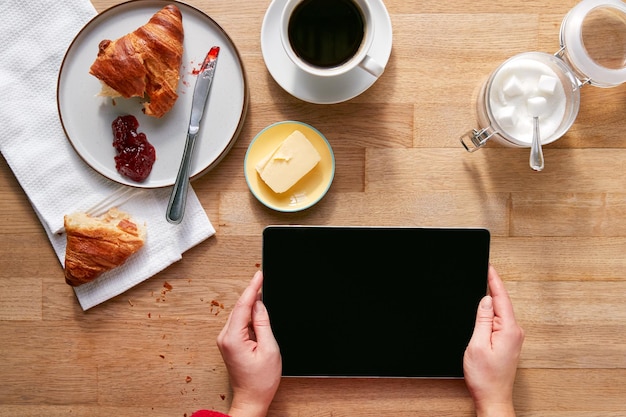 Overhead piatto laici di donna con tavoletta digitale sul tavolo apparecchiato per la colazione con croissant e caffè