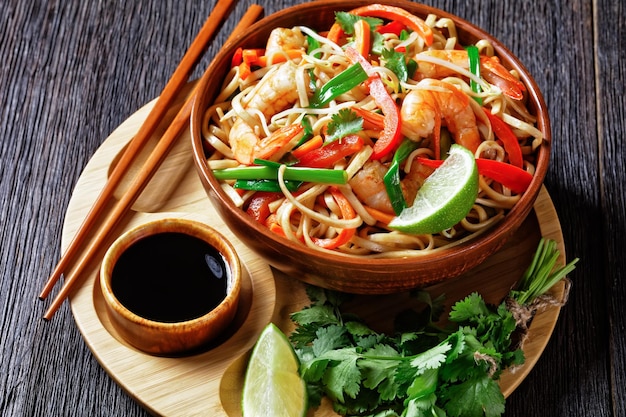 Pad thai noodles saltati in padella con gamberi, carote, peperoncino, coriandolo e spicchi di lime in una ciotola su un vassoio di bambù con salsa di soia e bacchette