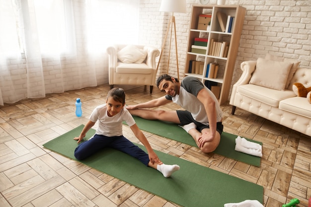 Padre And Daughter Stretch Legs On Gym Carpet.