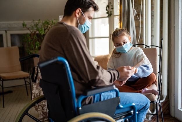 Padre con disabilità in sedia a rotelle che utilizza tablet a casa con bambino indossando maschere. Concetto di coronavirus della tecnologia dell'educazione delle persone