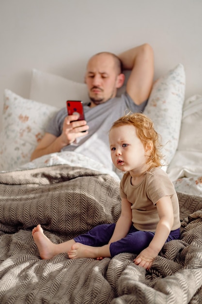 Padre del giovane che si stende sul letto facendo uso del cellulare con il suo bambino del bambino che si siede in primo piano a