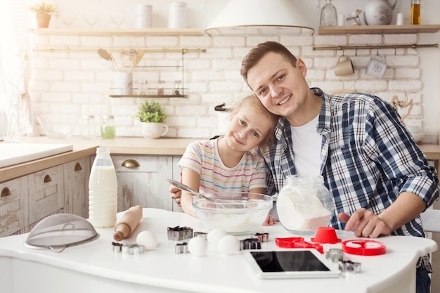 Padre e figlia che cuociono i biscotti. Bambina e suo padre che leggono la ricetta sulla tavoletta digitale in cucina a casa. Festa del papà, famiglia amorevole, concetto di vacanza