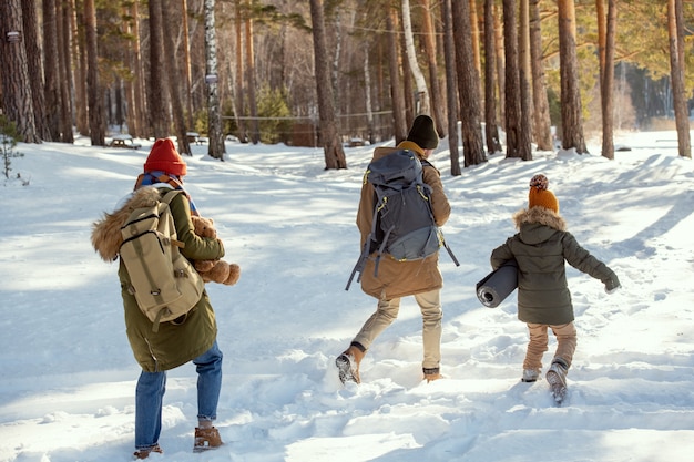 Padre e figlia in abiti invernali caldi che si divertono nel cumulo di neve mentre si spostano verso la casa di campagna, madre con lo zaino che li segue