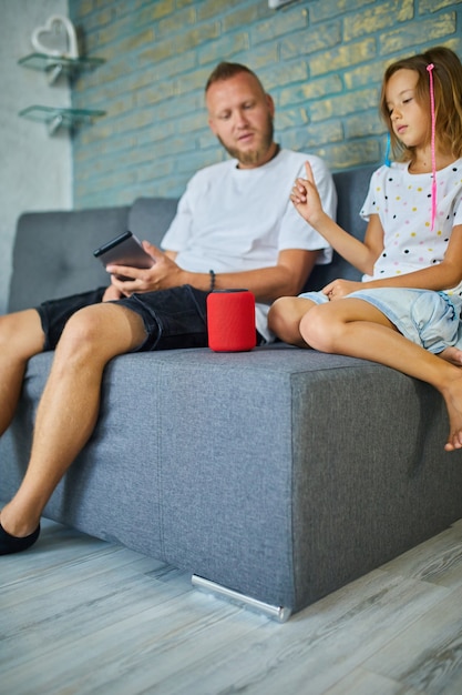 Padre e figlia provano a parlare con un altoparlante wireless intelligente, controllando i dispositivi domestici con comandi vocali, seduti sul divano di casa, concetto di casa intelligente