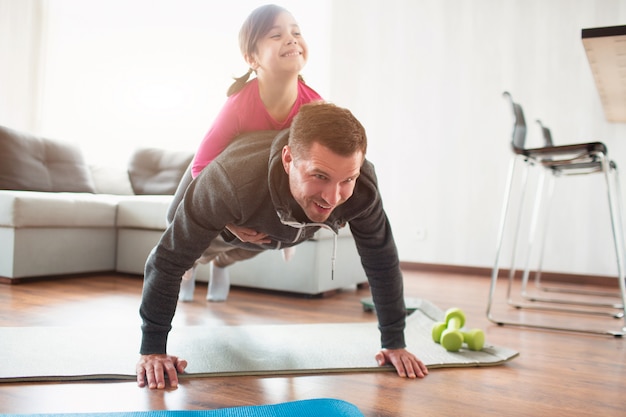 Padre e figlia si allenano a casa. Allenamento nell'appartamento. Sport in condizioni di casa. Fanno la tavola facendo. la figlia è salita sul papà. Si sta alzando da terra.