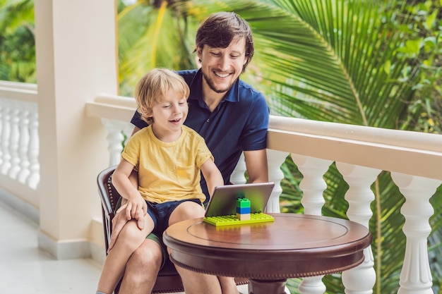 Padre e figlio ai tropici che parlano con amici e familiari in videochiamata utilizzando un tablet e cuffie wireless seduti sulla terrazza.