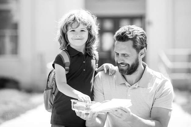 Padre e figlio americani che camminano attraverso il parco scolastico Scolaro e genitore in camicia con scatola per il pranzo