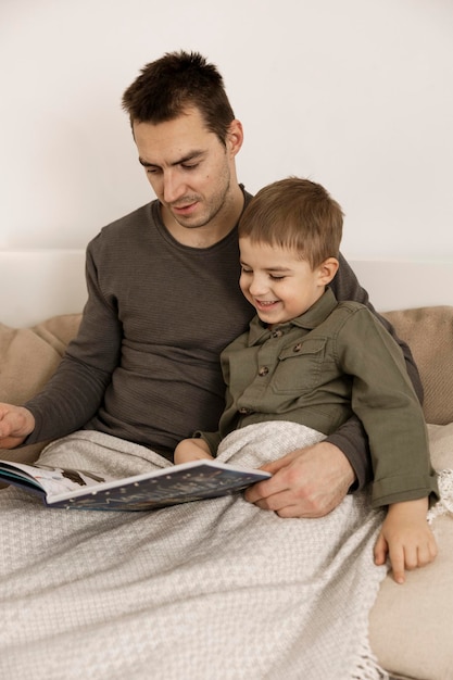 Padre e figlio che leggono un libro sul letto a casa. Giovane uomo attraente e ragazzino che riposano in camera da letto. Colori naturali della terra. Ambiente accogliente. Il padre legge una fiaba per suo figlio.