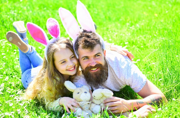 Padre e figlio con simpatiche orecchie da coniglio sdraiato sul prato Uomo con barba e bambino carino sdraiato sull'erba con un coniglietto in mano Papà e ragazza giocano nel giardino di primavera in una giornata di sole Concetto di festa di famiglia