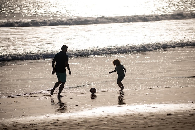 Padre e figlio giocano a calcio o a calcio sulla spiaggia durante le vacanze estive in famiglia Papà e figlio si divertono all'aperto Papà con bambino che gioca in una giornata estiva silhouette al tramonto