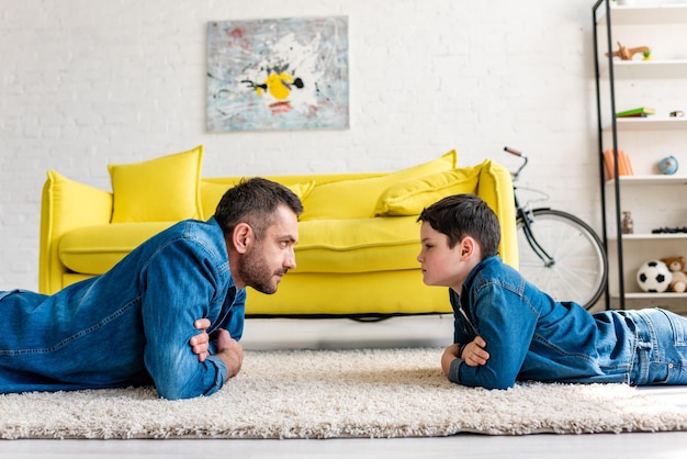 Padre e figlio in denim con le braccia incrociate sdraiati sul tappeto e guardandosi l'un l'altro a casa