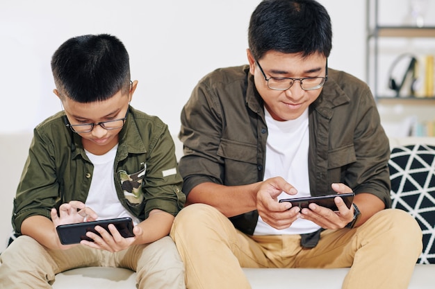 Padre e figlio vietnamiti con gli occhiali seduti sul divano e giocare con gli smartphone