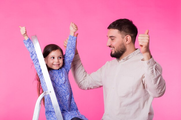 Padre e la sua piccola figlia con i pollici in su sopra il muro rosa. L'uomo adulto e la neonata sono felici.