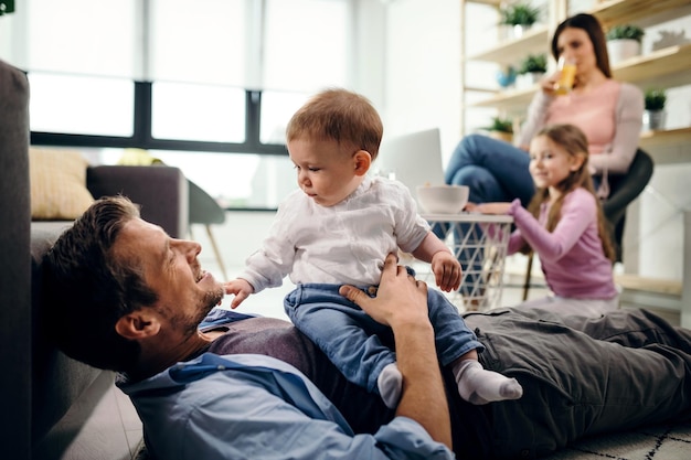 Padre felice che si rilassa con il suo bambino sul pavimento a casa mentre madre e figlia sono sullo sfondo