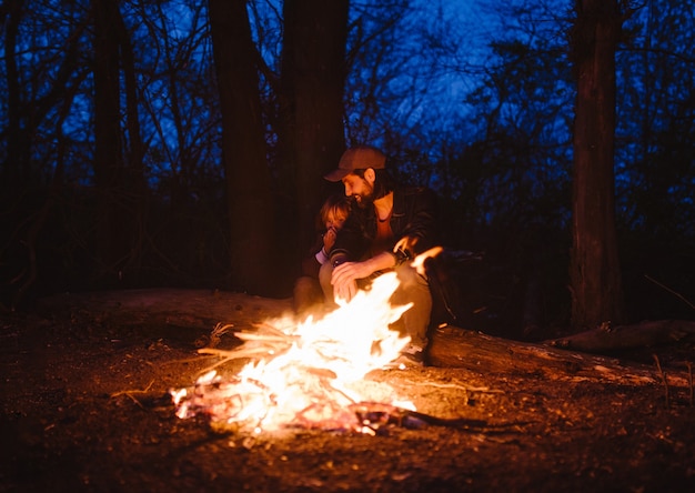 Padre felice e il suo figlioletto seduti insieme sui tronchi davanti a un fuoco in un'escursione nella foresta di notte. .