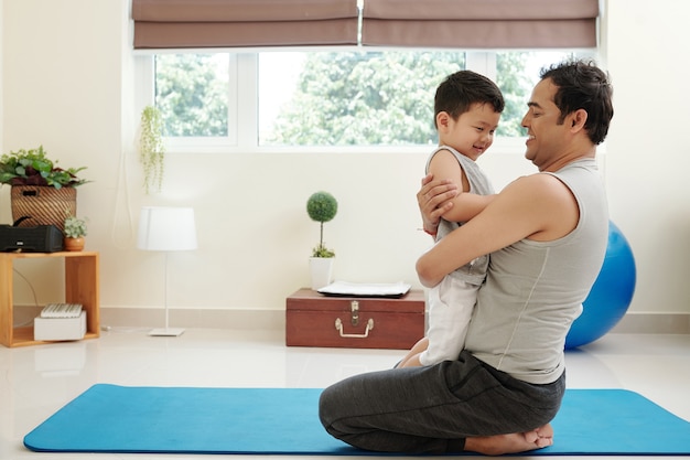 Padre felice seduto sul tappetino da yoga e abbracciando il suo piccolo figlio sorridente