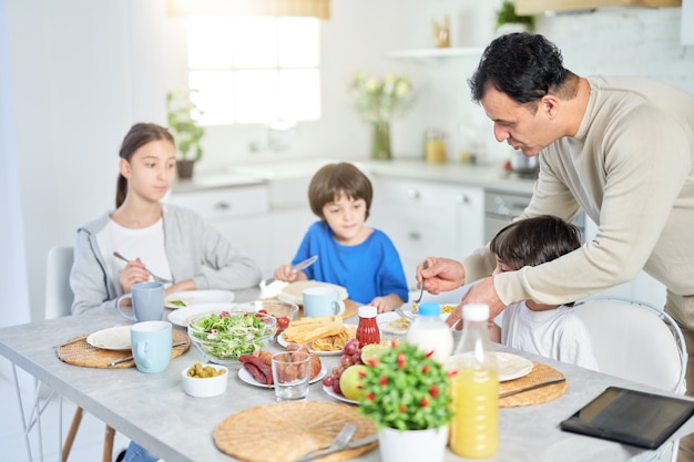 Padre latino premuroso che serve i suoi adorabili bambini mentre fanno colazione insieme a casa. Infanzia, genitorialità, concetto di cucina latina