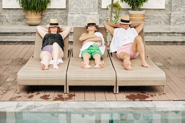 Padre, madre e figlio che si coprono il viso con il cappello quando prendono il sole sulle sdraio a bordo piscina