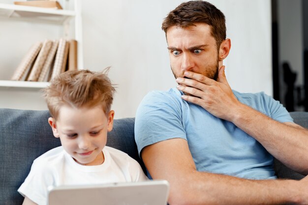 Padre preoccupato che dà una occhiata a ciò che suo figlio sta guardando sulla tavoletta digitale seduto sul divano di casa