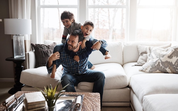 Padre ragazzi e bambini che giocano in soggiorno divertenti e spensierati con energia d'amore e felici a casa di famiglia L'uomo gioca con i bambini sul divano pazzo e giocoso con le spalle ed eccitato dalle risate