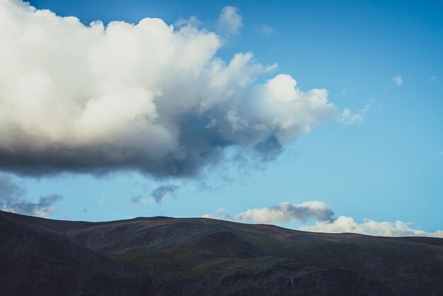 Paesaggio alpino minimalista con silhouette di montagna sotto il cielo nuvoloso con grande nuvola. Montagna scura e cielo chiaro. Fondo minimo della natura con la siluetta della montagna sotto la grande nuvola in cielo blu.