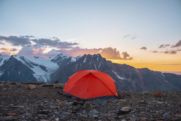 Paesaggio alpino panoramico con tenda ad altitudini molto elevate con vista su grandi montagne nel cielo arancione dell'alba Tenda arancione vivida con splendida vista sull'alta catena montuosa sotto il cielo nuvoloso nei colori del tramonto