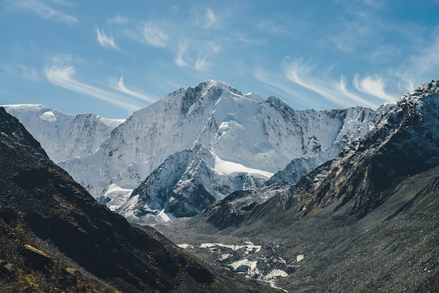 Paesaggio alpino soleggiato con alta montagna innevata con cima a punta e ghiacciaio tra rocce nere sotto i cirri nel cielo. Grandi montagne coperte di neve al sole. Picco appuntito bianco-neve alla luce del sole.