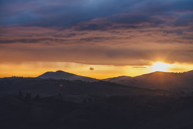 Paesaggio atmosferico con sagome di montagne con alberi sullo sfondo del cielo dell'alba con cerchio solare e raggi solari arancioni. Paesaggi naturali colorati con tramonto o alba di colori illuminanti.