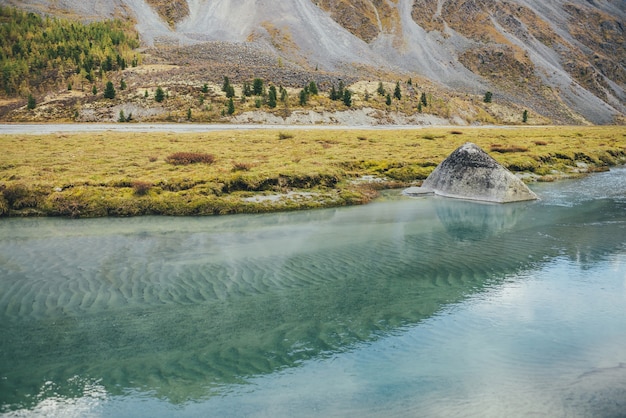 Paesaggio autunnale dorato con grande pietra nel torrente di montagna con vista sulla collina della foresta. Bellissimo grande masso nel flusso di acque turchesi in autunno. Fondale sabbioso verde ondulato in acqua trasparente.