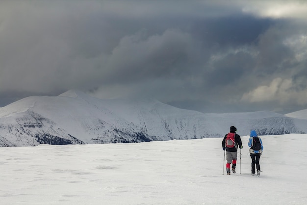 Paesaggio di montagna invernale. Vista posteriore di viaggiatori escursionisti turistici con zaini sul campo nevoso che camminano verso la montagna lontana su sfondo nuvoloso cielo tempestoso blu scuro copia spazio sulla fredda giornata invernale.