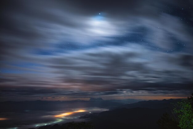 Paesaggio di nuvoloso che soffia attraverso il cielo blu con stelle e chiaro di luna sul picco di montagna nel parco nazionale di notte