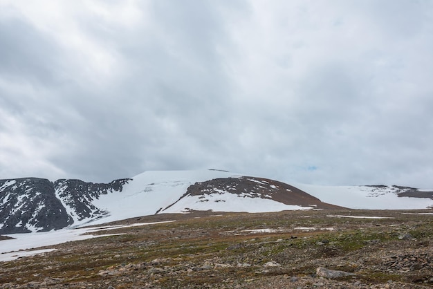 Paesaggio drammatico con campo di pietra e grande cupola di montagna innevata sotto il cielo nuvoloso grigio Scenario suggestivo con alta montagna di neve a forma di cupola al centro con tempo nuvoloso Coperto in montagna