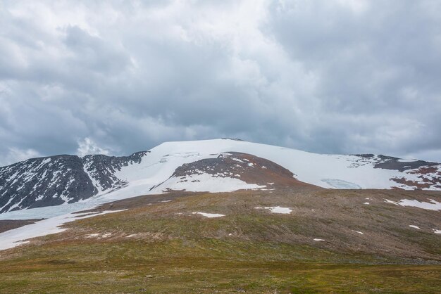 Paesaggio drammatico con grande cupola di montagna innevata alla luce del sole sotto il cielo nuvoloso Scenario colorato con alta montagna di neve illuminata dal sole a forma di cupola al centro con tempo variabile Vista panoramica sulle montagne