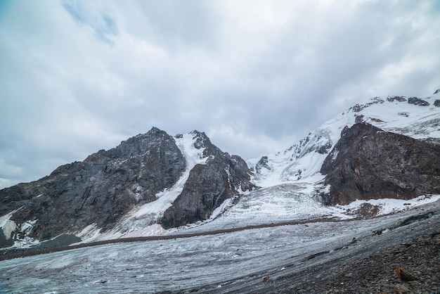 Paesaggio drammatico con lingua di ghiacciaio e cascata di ghiaccio su una grande catena montuosa innevata con picco roccioso sotto un cielo grigio e nuvoloso lungo ghiacciaio ad alta quota paesaggio cupo in montagna in nuvoloso