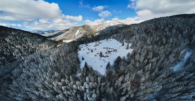 Paesaggio invernale aereo con piccole case rurali tra foreste innevate in montagne fredde.