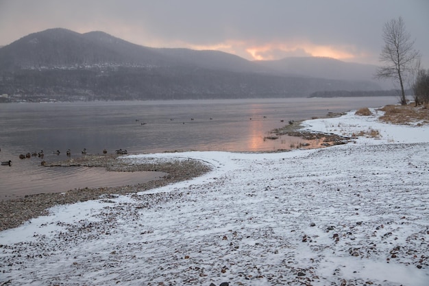 Paesaggio invernale al tramonto - fiume, ghiaccio sulla riva, montagne sullo sfondo